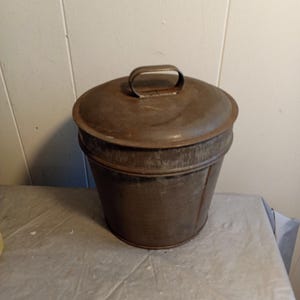 Könnte beinhalten: Ein alter Metallbehälter mit Deckel und Griff. Der Eimer hat eine dunkle, verwitterte braune Farbe und weist Gebrauchsspuren auf. Der Deckel hat einen kleinen, gewölbten Griff zum einfachen Anheben. Der Eimer steht auf einer hellen Oberfläche.