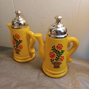 May include: Two yellow ceramic teapots with silver lids. Each teapot features a floral design with red flowers and green leaves in a basket. The teapots have a handle and a spout.