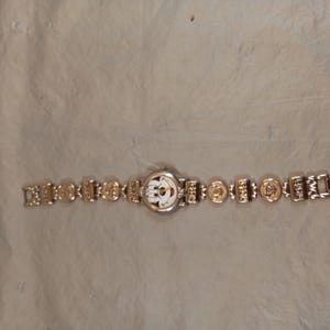 May include: A silver-tone wristwatch featuring a round white face with a colourful cartoon character graphic. The watch band is composed of alternating rectangular and circular metal links, each embossed with small detailed designs and patterns.