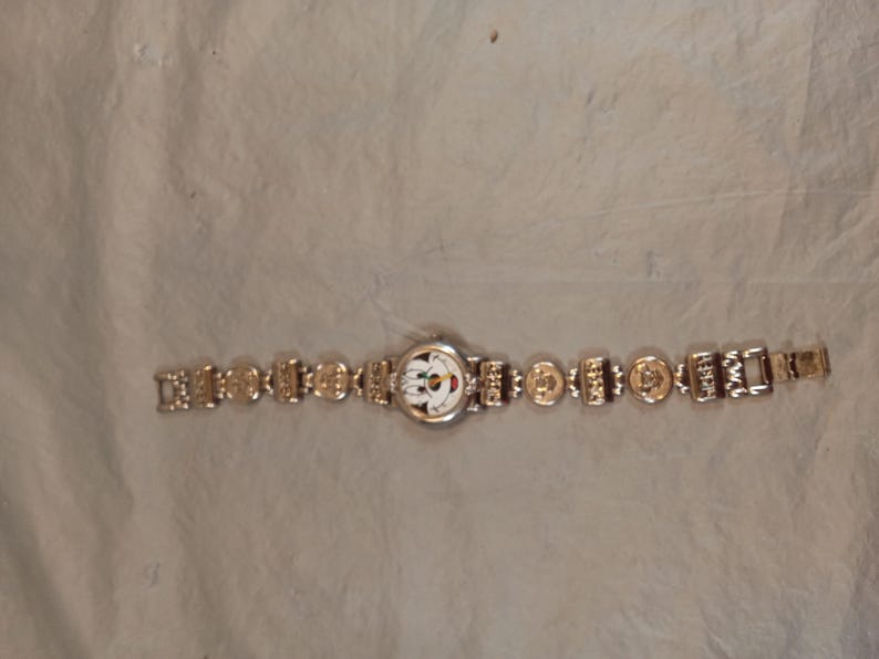 May include: A silver-tone wristwatch featuring a round white face with a colourful cartoon character graphic. The watch band is composed of alternating rectangular and circular metal links, each embossed with small detailed designs and patterns.