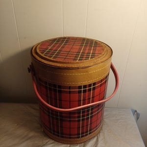 May include: A vintage round hat box with a red and black plaid pattern. The box has a tan rim and a pink handle. The lid also features the plaid design. The box is made of cardboard or a similar material.