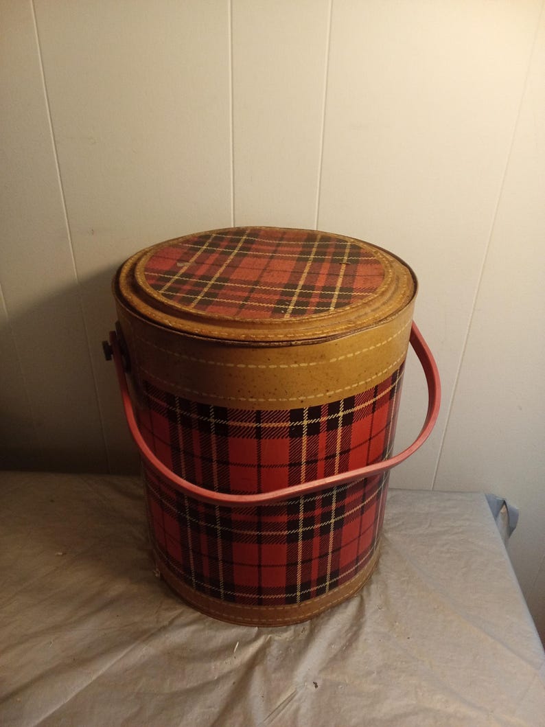May include: A vintage round hat box with a red and black plaid pattern. The box has a tan rim and a pink handle. The lid also features the plaid design. The box is made of cardboard or a similar material.
