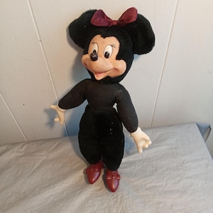 May include: A vintage Minnie Mouse plush doll. The doll has a black body, black ears, and a burgundy bow. The face is tan with black eyes and a black nose. The doll is wearing red shoes and white gloves.