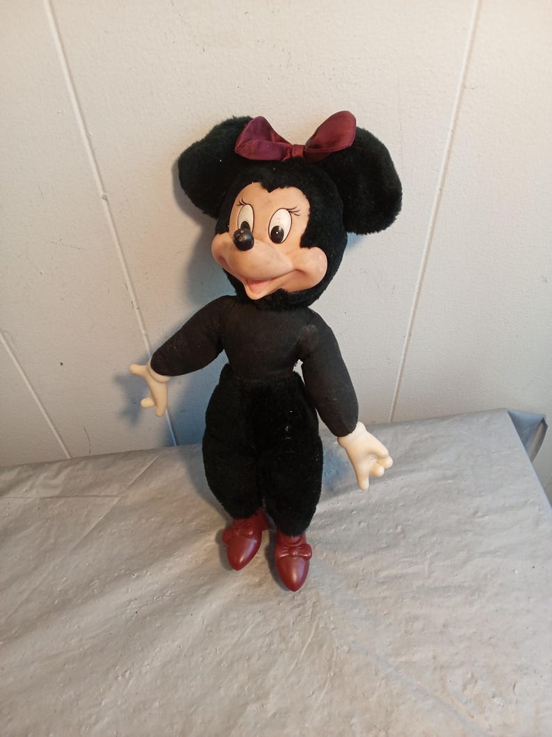 May include: A vintage Minnie Mouse plush doll. The doll has a black body, black ears, and a burgundy bow. The face is tan with black eyes and a black nose. The doll is wearing red shoes and white gloves.