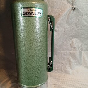 May include: A tall, cylindrical green vacuum flask with a textured, hammered finish. It features a side-mounted green plastic handle with metal hinges and a silver-colored base. A white label on the front displays the text Aladdin STANLEY.