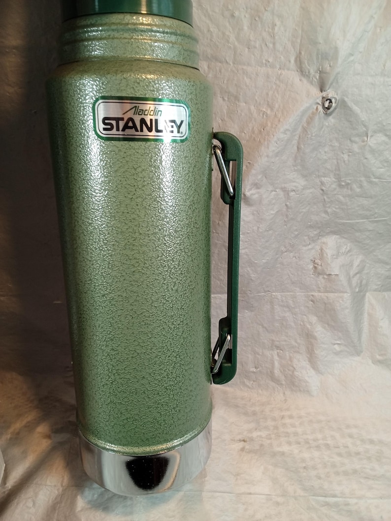 May include: A tall, cylindrical green vacuum flask with a textured, hammered finish. It features a side-mounted green plastic handle with metal hinges and a silver-colored base. A white label on the front displays the text Aladdin STANLEY.