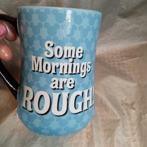 Pu&ograve; includere: Una tazza di ceramica azzurra con un manico nero. La tazza presenta un motivo di sagome bianche di Topolino e il testo "Some Mornings are ROUGH!" in lettere bianche.