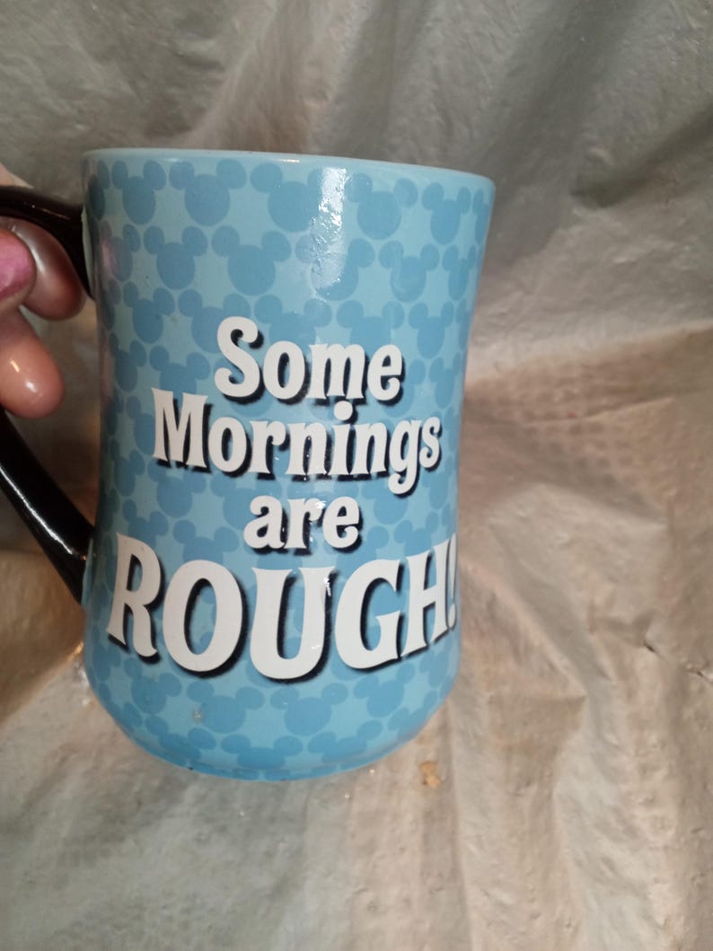 Pu&ograve; includere: Una tazza di ceramica azzurra con un manico nero. La tazza presenta un motivo di sagome bianche di Topolino e il testo "Some Mornings are ROUGH!" in lettere bianche.