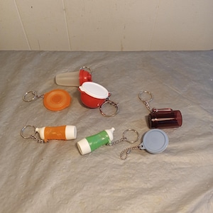 May include: A selection of miniature keyrings in various shapes and colours. Includes a small orange disc, a red container with a white lid, and cylindrical keyrings in orange, green, and brown. Each keyring has a metal ring and chain.