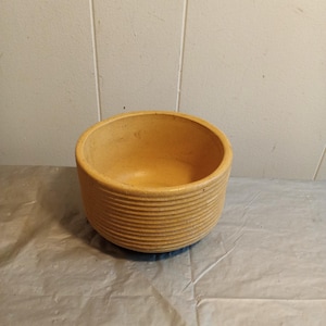 Op de afbeelding: Een ronde, lichtbruine keramische kom met horizontale ribbels aan de buitenkant. De kom heeft een brede opening en een gladde binnenkant. De kom staat op een lichtgekleurd oppervlak.