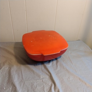 Puede incluir: Una fuente de horno de vidrio roja con tapa. La fuente es redonda y tiene un borde ligeramente elevado. La tapa también es redonda y tiene una manija en la parte superior.