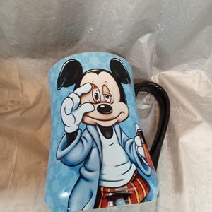 Pu&ograve; includere: Una tazza in ceramica con un'illustrazione a cartoni animati di Topolino. La tazza ha uno sfondo azzurro chiaro con Topolino con un cappotto blu e pantaloni a righe rosse e gialle. Il manico &egrave; nero.