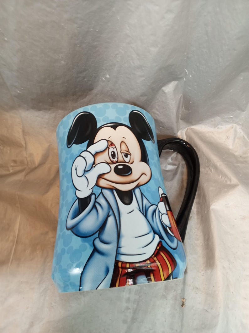 Pu&ograve; includere: Una tazza in ceramica con un'illustrazione a cartoni animati di Topolino. La tazza ha uno sfondo azzurro chiaro con Topolino con un cappotto blu e pantaloni a righe rosse e gialle. Il manico &egrave; nero.