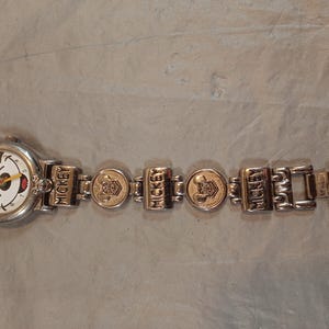 May include: A silver-tone analogue wristwatch featuring a white dial with a Mickey Mouse graphic, black numerals, and colourful hands. The metal link bracelet displays the name MICKEY and embossed character head icons on alternating rectangular and round links.