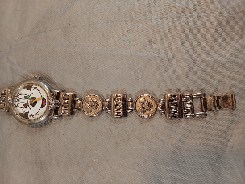 May include: A silver-tone analogue wristwatch featuring a white dial with a Mickey Mouse graphic, black numerals, and colourful hands. The metal link bracelet displays the name MICKEY and embossed character head icons on alternating rectangular and round links.