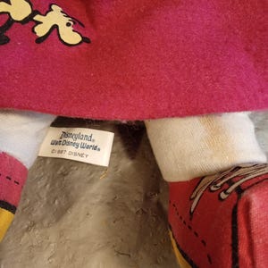 May include: Close-up of a vintage doll with a red felt dress and white arms. The doll's shoes are red with yellow soles. The tag reads "Disneyland, Walt Disney World, &copy;1987 Disney".