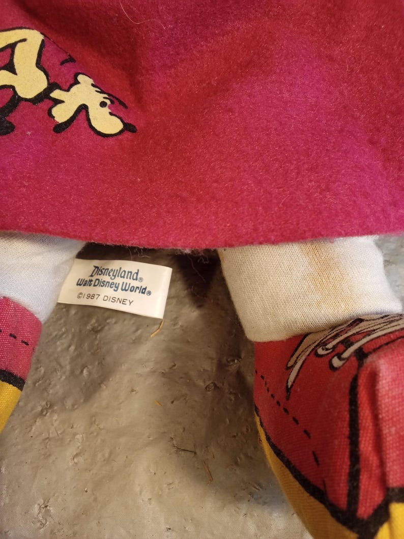 May include: Close-up of a vintage doll with a red felt dress and white arms. The doll's shoes are red with yellow soles. The tag reads "Disneyland, Walt Disney World, &copy;1987 Disney".