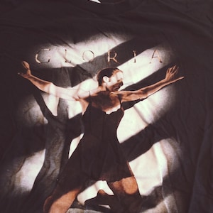 May include: Black t-shirt with a white graphic of a woman in a black dress with the word "GLORIA" printed in gold letters.