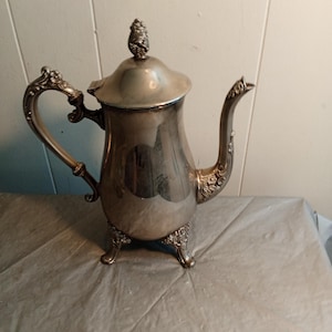 Puede incluir: Una tetera plateada con detalles ornamentados. La tetera tiene un asa curvada, un pico y una tapa con un adorno decorativo. Se apoya sobre tres pequeños pies. La tetera es probablemente de metal y tiene una superficie reflectante.
