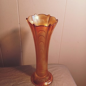 May include: An amber-colored glass vase with a flared, ruffled rim. The vase has a tall, slender body with vertical ridges and a rounded base. The glass has an iridescent sheen, catching the light.