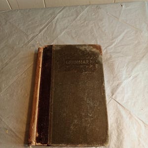 May include: An antique book titled "Grammar" with a worn, textured cover. The book has a dark brown spine and a faded green cover, showing signs of age and use. The edges of the pages are visible, and the book appears to be well-loved.