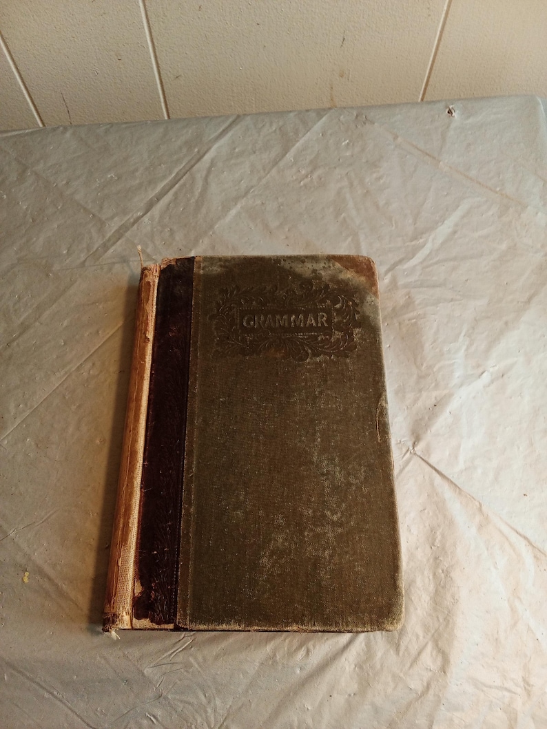 May include: An antique book titled "Grammar" with a worn, textured cover. The book has a dark brown spine and a faded green cover, showing signs of age and use. The edges of the pages are visible, and the book appears to be well-loved.