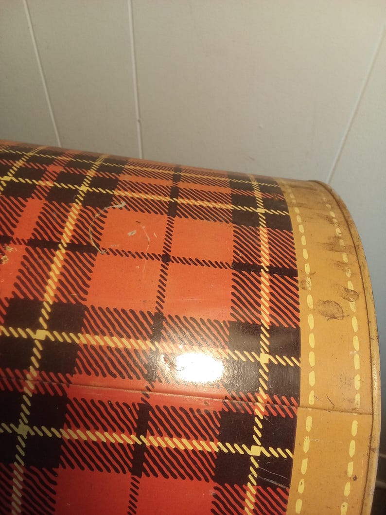 May include: A vintage metal container with a red, black, and yellow plaid pattern. The container has a rounded shape and a tan border with a dotted design. The plaid design is a classic pattern.