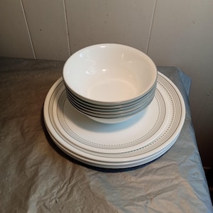 Puede incluir: Un juego de cuencos y platos de cerámica blanca. Los cuencos están apilados sobre los platos. Los platos y cuencos tienen un diseño de borde gris punteado y rayado. La vajilla está sobre una superficie gris.