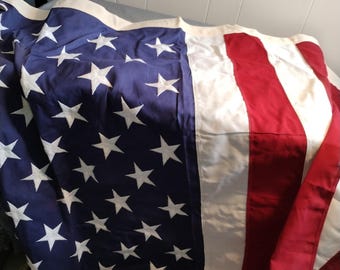 Può includere: Una grande bandiera americana con stelle bianche su un campo blu e strisce rosse e bianche. La bandiera è piegata e sembra essere fatta di un tessuto resistente.