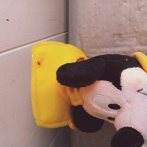 Puede incluir: Un peluche de Mickey Mouse con orejas negras, cara rosa y atuendo amarillo. Solo se ve la cabeza y parte del cuerpo. El fondo es una pared de color claro.