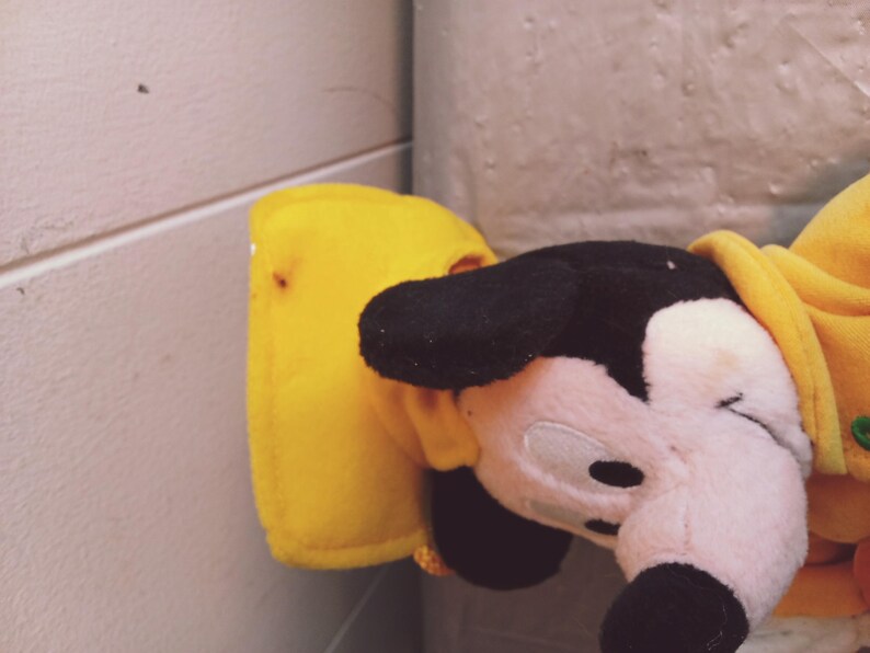 Puede incluir: Un peluche de Mickey Mouse con orejas negras, cara rosa y atuendo amarillo. Solo se ve la cabeza y parte del cuerpo. El fondo es una pared de color claro.