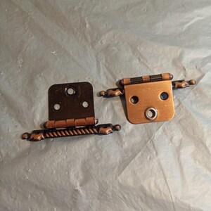May include: Two antique-style cabinet hinges. One is a dark bronze color, the other is copper. Both have decorative rope-like detailing and rounded ends. The hinges are designed for overlay cabinet doors.