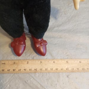 May include: A doll's legs and feet are visible, wearing dark green pants and red shoes. The shoes have a bow detail. A wooden ruler with inch measurements is in the foreground.