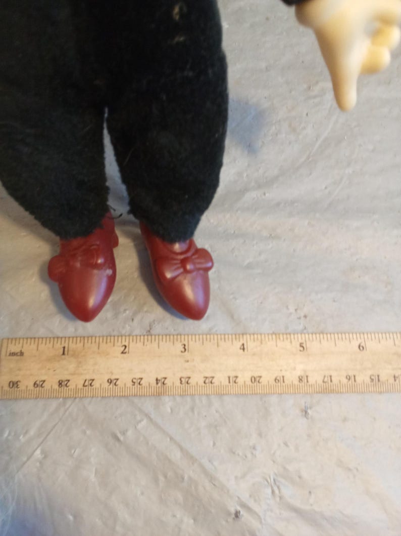 May include: A doll's legs and feet are visible, wearing dark green pants and red shoes. The shoes have a bow detail. A wooden ruler with inch measurements is in the foreground.