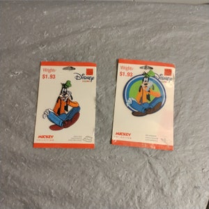 Op de afbeelding: Twee strijkbare patches met Goofy van Disney. De patches bevinden zich op witte kaarten met rode en blauwe randen. De prijs is $ 1,93. De tekst op de kaarten luidt "Wrights", "Disney Home", "Mickey Collection", "Iron On", "Patch/Adhesive", "Auto For Clothes", "Self-Adhesive", "Autocollante", "Autoadhesiva".