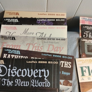 Pode incluir: Uma pilha de livros de bolso coloridos com o t&iacute;tulo "Leisure Arts" e v&aacute;rios t&iacute;tulos de projetos de artesanato, incluindo "More All", "From This Day", "Kathie's Dreams", "Discovery The New World" e "Flowers About The House".