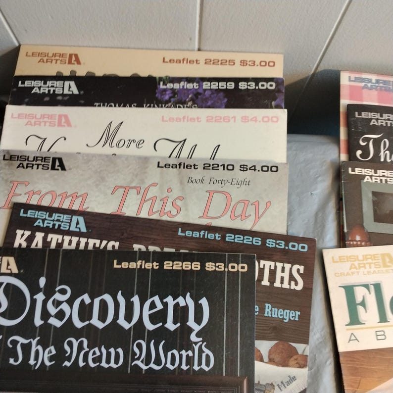 Pode incluir: Uma pilha de livros de bolso coloridos com o t&iacute;tulo "Leisure Arts" e v&aacute;rios t&iacute;tulos de projetos de artesanato, incluindo "More All", "From This Day", "Kathie's Dreams", "Discovery The New World" e "Flowers About The House".