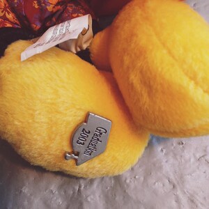 Puede incluir: Primer plano de un juguete de peluche amarillo con una etiqueta que dice "Graduation 2003". El juguete est&aacute; hecho de un material suave y texturizado. El fondo incluye un vistazo de tela estampada.