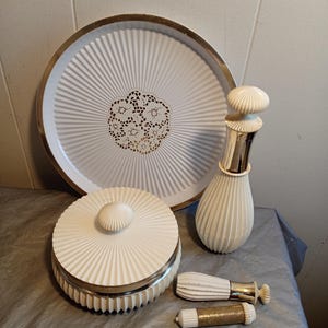 May include: A vintage vanity set featuring a white tray with a gold rim and a gold filigree design in the center. The set includes a jar, a perfume bottle, and two cosmetic applicators, all in white with silver accents.