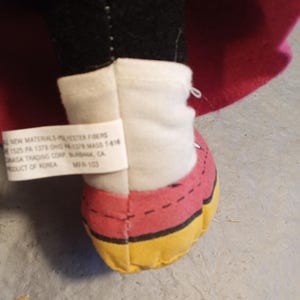 May include: A close-up of a plush toy's foot, featuring a red shoe with yellow trim and a white ankle cuff. The toy's black leg and a pink cape are visible. A tag with text is attached to the cuff.