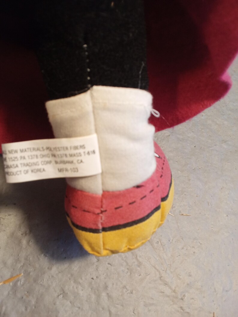 May include: A close-up of a plush toy's foot, featuring a red shoe with yellow trim and a white ankle cuff. The toy's black leg and a pink cape are visible. A tag with text is attached to the cuff.