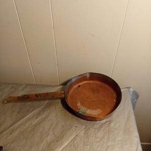 May include: A well-worn copper frying pan with a long, rectangular handle. The pan's interior shows signs of use, with a slightly discolored surface. The handle appears to be made of the same material as the pan, with a rustic, aged appearance.