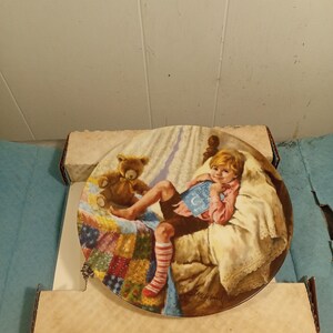 Könnte beinhalten: Dekorative Platte mit einem Kind, das im Bett mit einem Teddybären liest. Die Platte zeigt eine bunte Steppdecke und den Text "Dear Diary". Das Kind trägt ein rosa Hemd und gestreifte Socken.