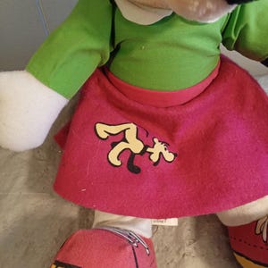 May include: A plush Minnie Mouse doll wearing a green top, a pink skirt with a cartoon graphic, and red shoes with yellow soles. The doll has a white collar and white gloves. The Disney tag is visible.
