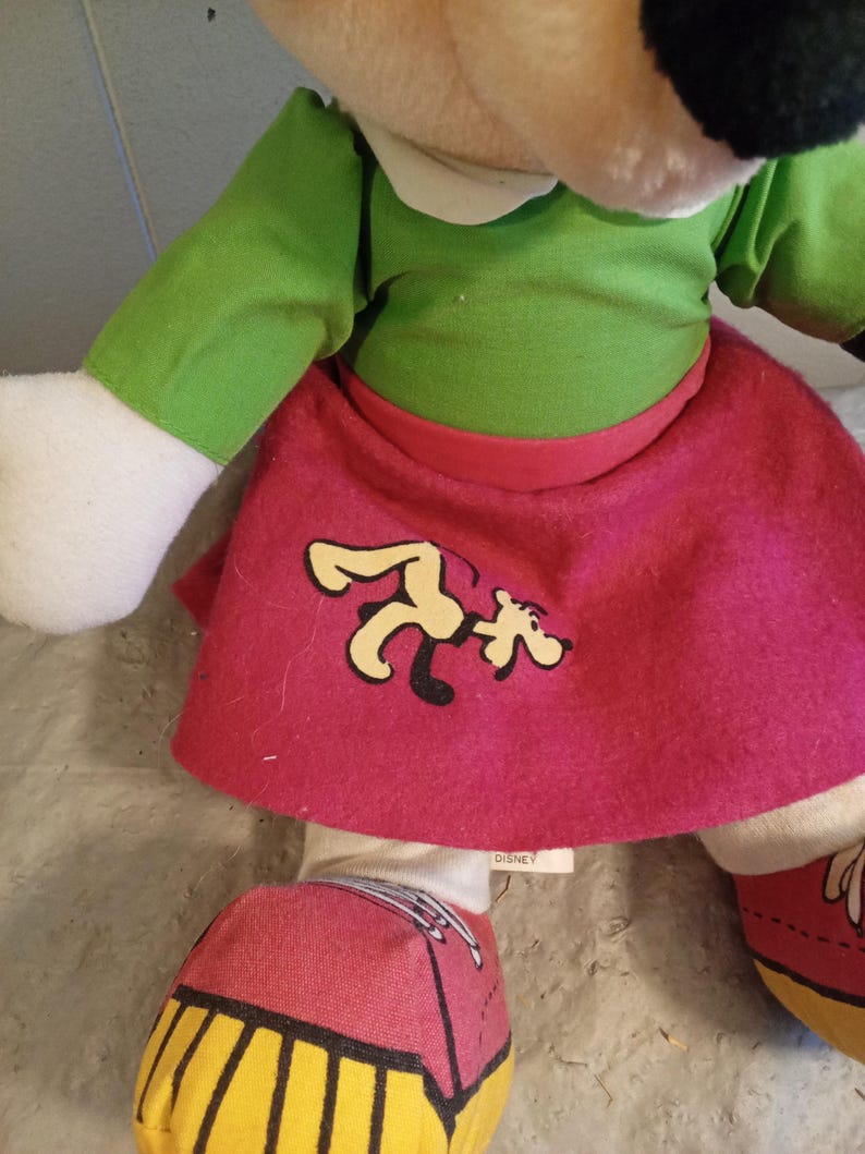 May include: A plush Minnie Mouse doll wearing a green top, a pink skirt with a cartoon graphic, and red shoes with yellow soles. The doll has a white collar and white gloves. The Disney tag is visible.
