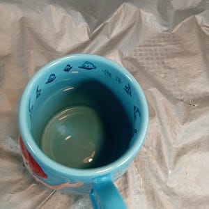 May include: A light blue ceramic mug featuring a mermaid design. The mug has a matching blue handle and a light blue interior. The rim of the mug is decorated with small blue sea creature illustrations.