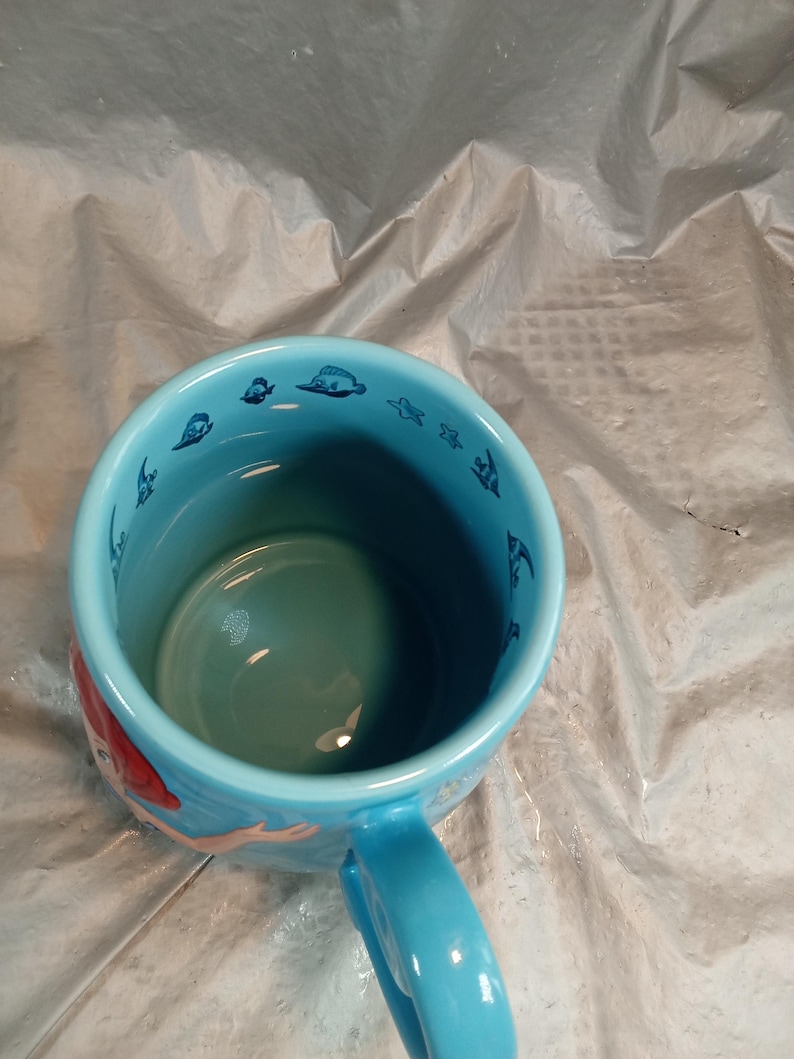 May include: A light blue ceramic mug featuring a mermaid design. The mug has a matching blue handle and a light blue interior. The rim of the mug is decorated with small blue sea creature illustrations.