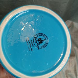May include: A turquoise ceramic mug with a white rim and a blue handle. The bottom of the mug features a Disney Parks logo and the text "Authentic Original" and "Made in Thailand".