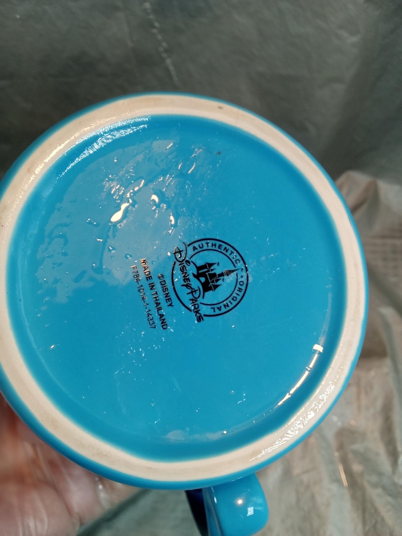 May include: A turquoise ceramic mug with a white rim and a blue handle. The bottom of the mug features a Disney Parks logo and the text "Authentic Original" and "Made in Thailand".