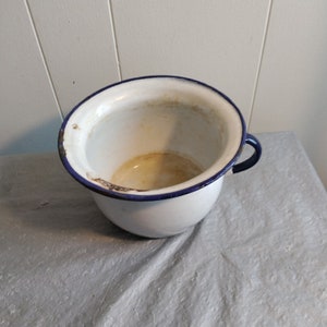 May include: White enamel chamber pot with a blue rim and a single handle. The pot is slightly dirty and has a few rust spots.